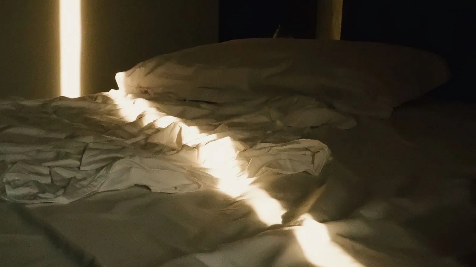 Una cama deshecha en una habitación a oscuras, con una franja de luz de la mañana